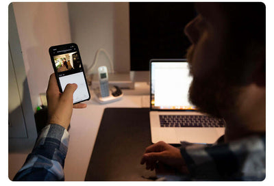 Person monitoring baby through Smart Camera Indoor Baby Monitor on smartphone beside a laptop in a dimly lit room.